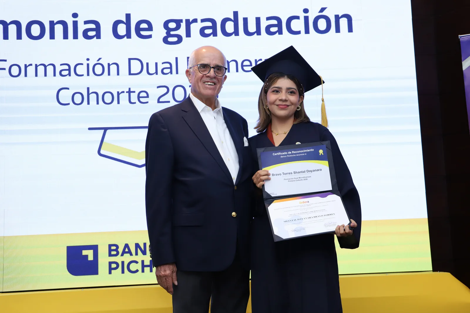 Una graduada de la primera cohorte muestra su certificado junto a un directivo de Banco Pichincha